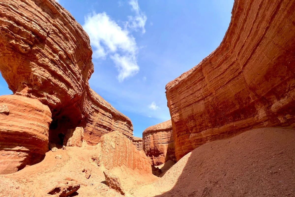 Czerwone, wyżłobione skały tworzą imponujący kanion z licznymi warstwami geologicznymi, kontrastującymi z błękitnym niebem i białymi obłokami.