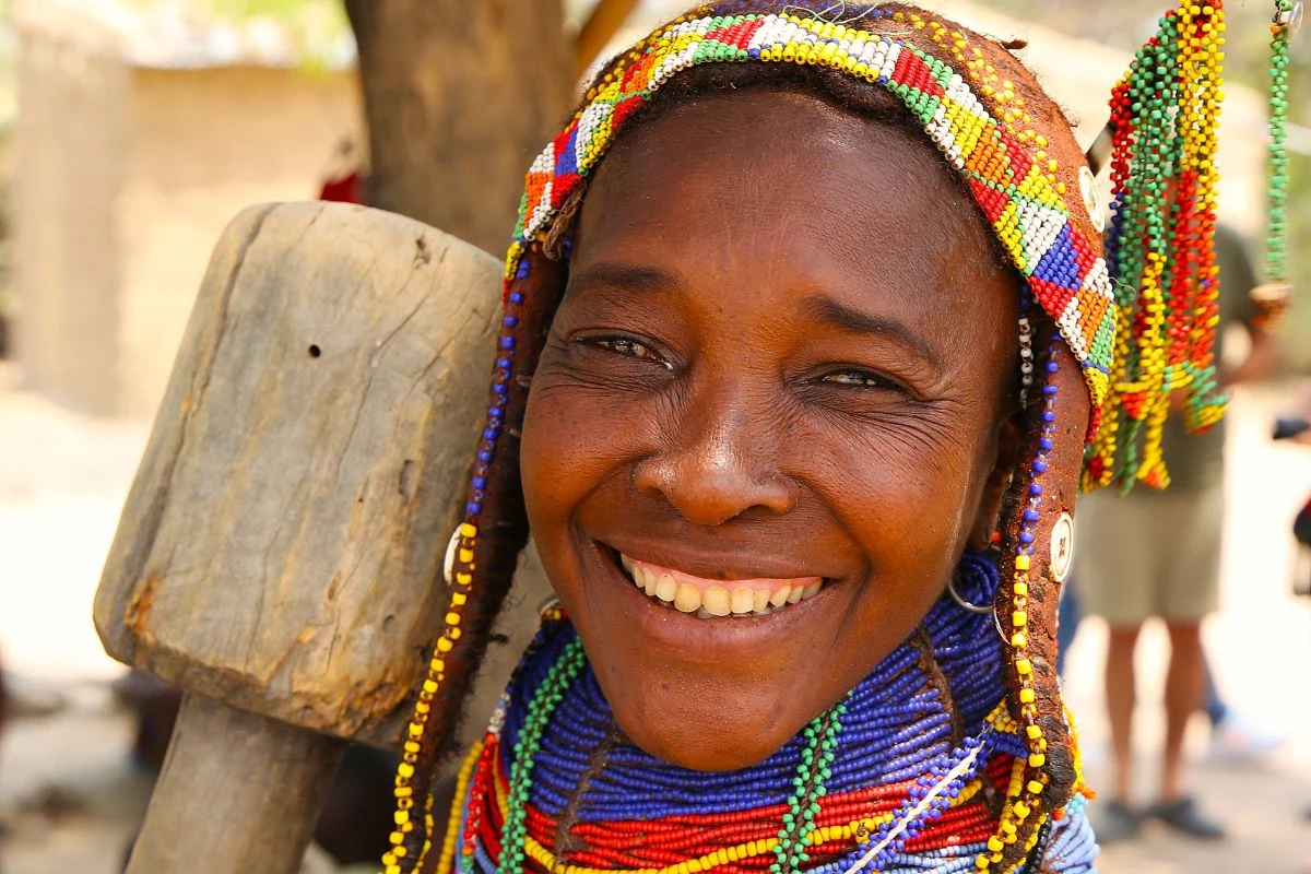 Uśmiechnięta kobieta w kolorowych koralikach i tradycyjnym stroju etnicznym, tło rozmyte, widać fragment drewnianego młotka lub narzędzia obok niej.