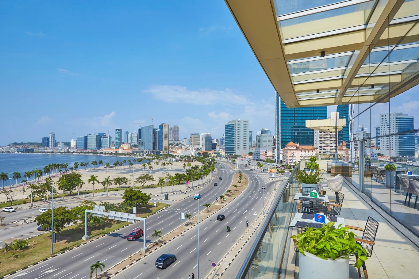 Widok z nowoczesnego tarasu na szeroką, nadmorską promenadę oraz rozległą miejską panoramę z wysokimi budynkami i palmami. Jasne, słoneczne niebo oraz pusta droga podkreślają klimat dużego miasta nad wodą.