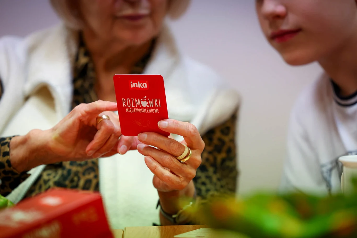 Starsza kobieta i młody chłopiec siedzą przy stole, trzymają czerwoną kartę z napisem 'Inka Rozmówki Międzypokoleniowe'. W tle widać rozmyte elementy i kartonik, a w centrum uwagi są dłonie kobiety z pierścionkami.