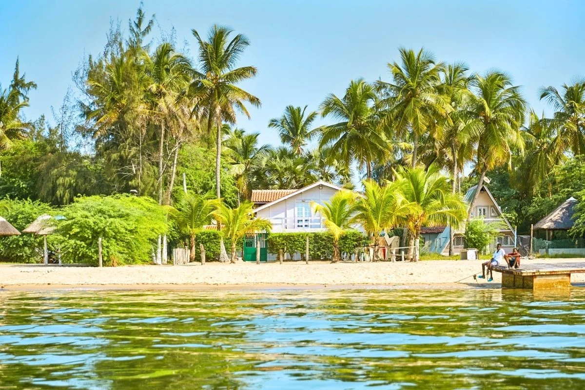 Plaża z jasnym piaskiem otoczona licznymi palmami, w tle kilka domów wypoczynkowych i gęsta roślinność tropikalna. Na brzegu widocznych jest kilka osób odpoczywających, a przed nimi spokojna tafla wody.