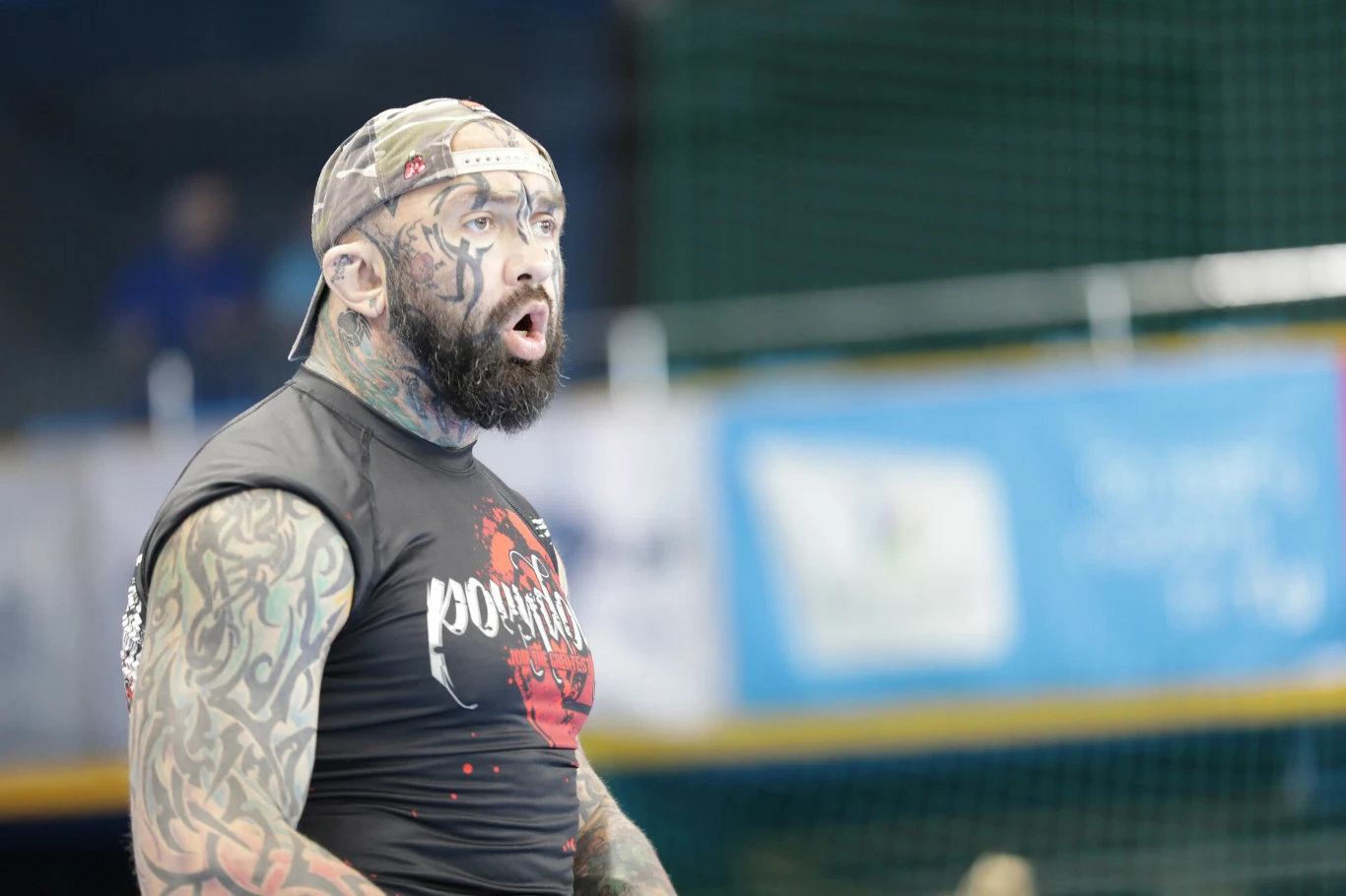 Mężczyzna z licznymi tatuażami na twarzy i ramionach, ubrany w czarną koszulkę z nadrukiem, noszący czapkę z daszkiem, stoi w hali sportowej z wyrazem zaskoczenia lub ekscytacji na twarzy.