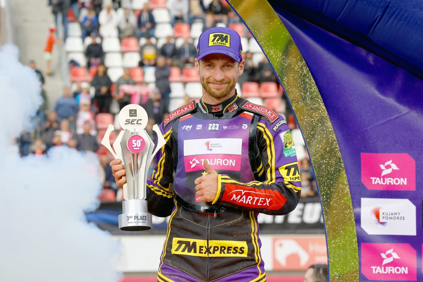 Kacper Woryna na podium Tauron SEC w Bydgoszczy 2025 Sportowiec w stroju motocyklowym trzyma w ręku puchar za zajęcie pierwszego miejsca, uśmiecha się do aparatu. W tle widoczne są trybuny z kibicami oraz kolorowe elementy dekoracji związanych ze sponsorem wydarzenia.