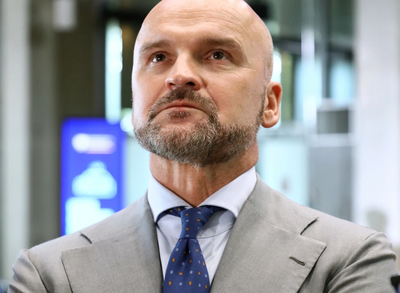 Rafał Brzoska oddaje w ręce użytkowników aplikacji InPost przyszłość polskiego modelu językowego Bielik LLM Mężczyzna łysy z brodą i krótkim zarostem, ubrany w szary garnitur, niebieską koszulę i niebieski krawat w drobny wzór, patrzący poważnie do góry. W tle nieostre biurowe wnętrze z niebieskim ekranem informacyjnym.