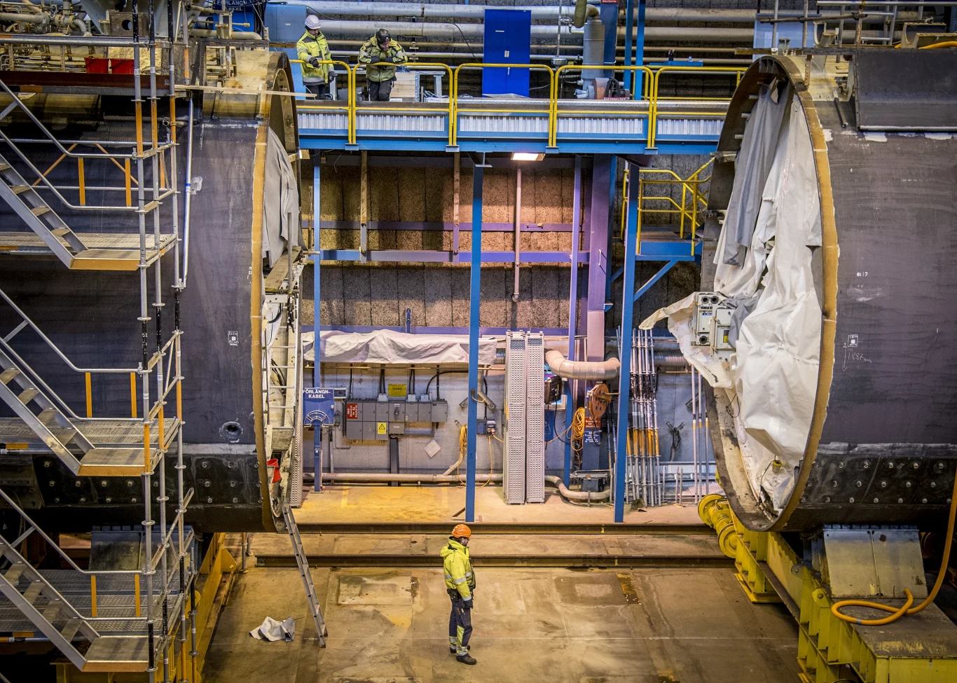 Wnętrze dużej hali przemysłowej z masywną maszyną budowlaną oraz pracownikami w odzieży ochronnej, którzy nadzorują postęp prac przy tunelu lub konstrukcji przemysłowej.