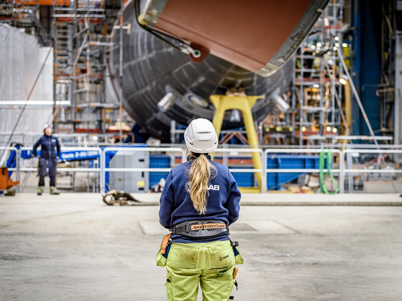 Kobieta w odzieży roboczej i kasku ochronnym stoi tyłem na hali przemysłowej, obserwując fragment dużego obiektu technicznego otoczonego rusztowaniami i sprzętem budowlanym.