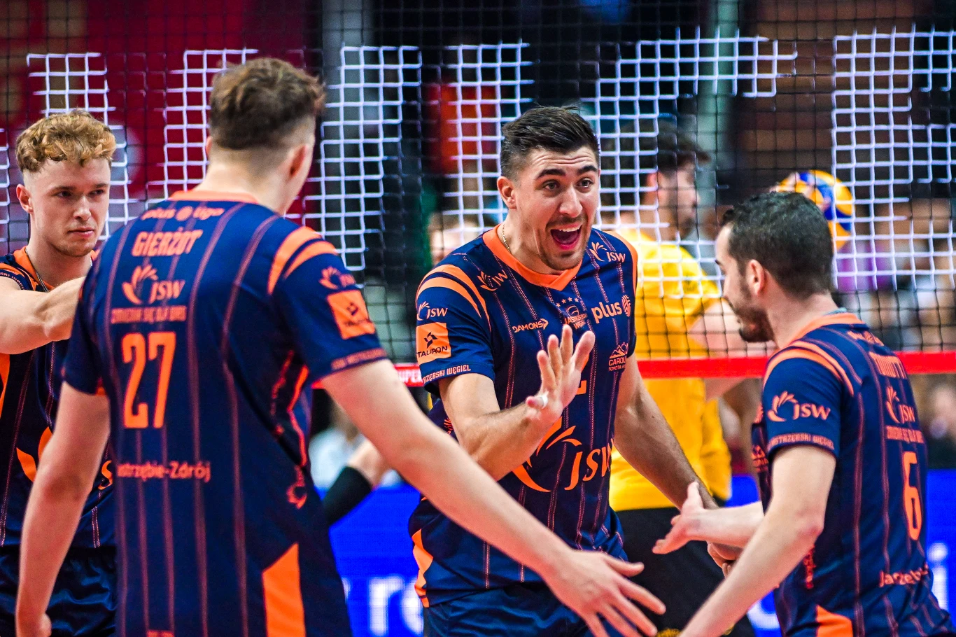 Siatkarze JSW Jastrzębskiego Węgla Grupa siatkarzy w niebiesko-pomarańczowych strojach drużynowych świętuje zdobycie punktu podczas meczu, ekscytacja i pozytywne emocje dominują na twarzach sportowców, w tle widać siatkę i fragment trybun.
