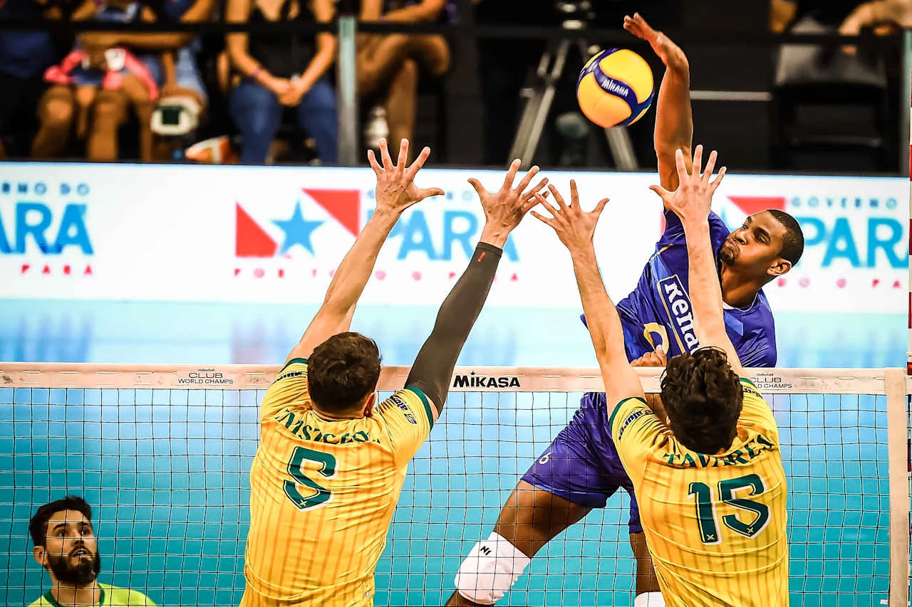 Zawodnik w niebieskim stroju atakuje piłkę do siatkówki nad siatką, próbując pokonać dwóch przeciwników w żółto-zielonych koszulkach, którzy wykonują blok. Na trybunach widoczna publiczność, w tle reklama zawodów.