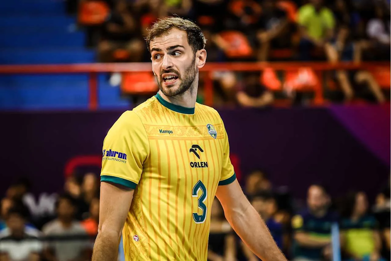 Aaron Russell, przyjmujący Warty Siatkarz w żółtej koszulce z numerem 3 podczas meczu siatkówki, na drugim planie widownia w rozmyciu oraz barwne trybuny.