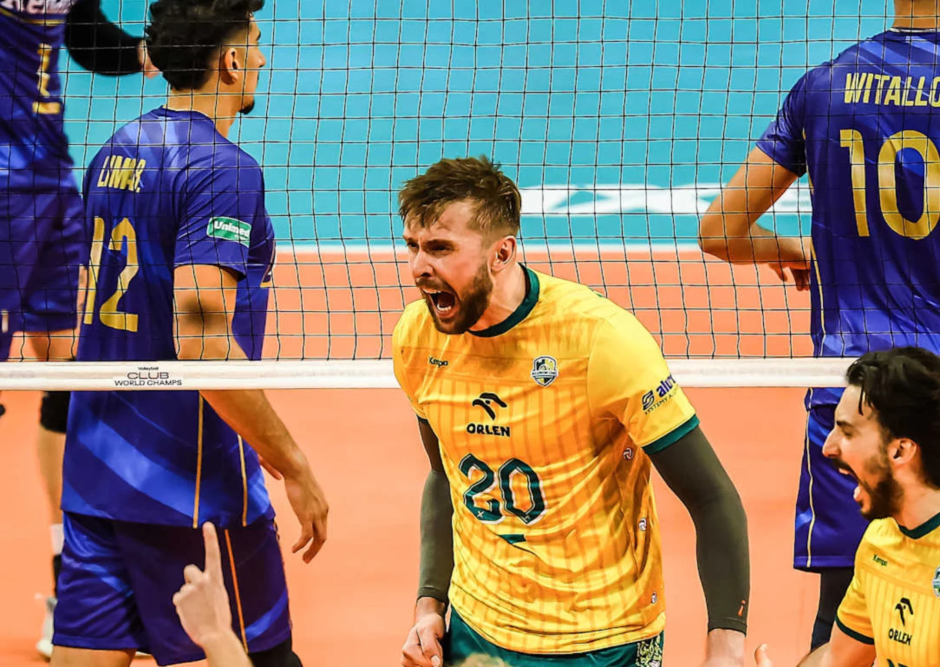 Zawodnik drużyny siatkarskiej w żółtym stroju wyraża silne emocje podczas rozgrywki, otoczony przez innych graczy w niebieskich koszulkach, na tle siatki na hali sportowej.