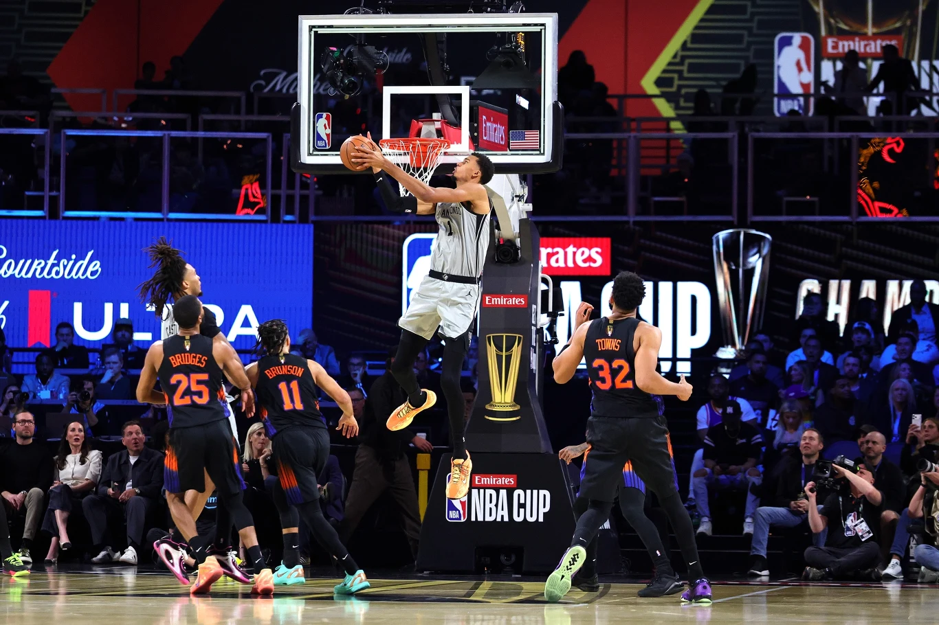 Koszykarz w białym stroju skacze do wsadu przy koszu, otoczony przez czterech obrońców w czarnych strojach na tle pełnej widowni. W tle widoczne banery sponsorów oraz napis NBA CUP.