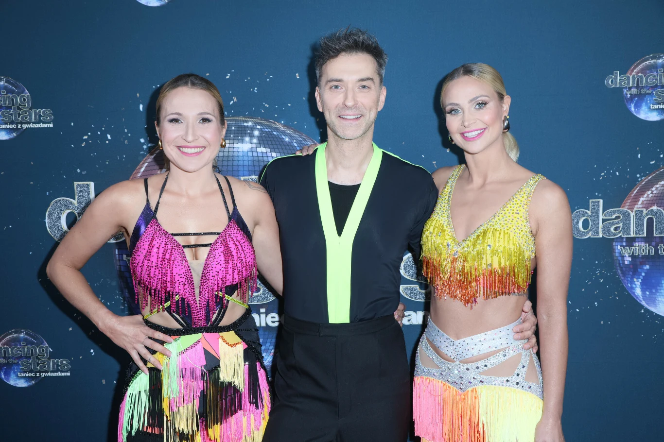 Trzy uśmiechnięte osoby w kolorowych, błyszczących strojach tanecznych stoją na tle z logo programu telewizyjnego. Atmosfera jest radosna, dominują barwne frędzle, cekiny oraz wyraziste kolory.