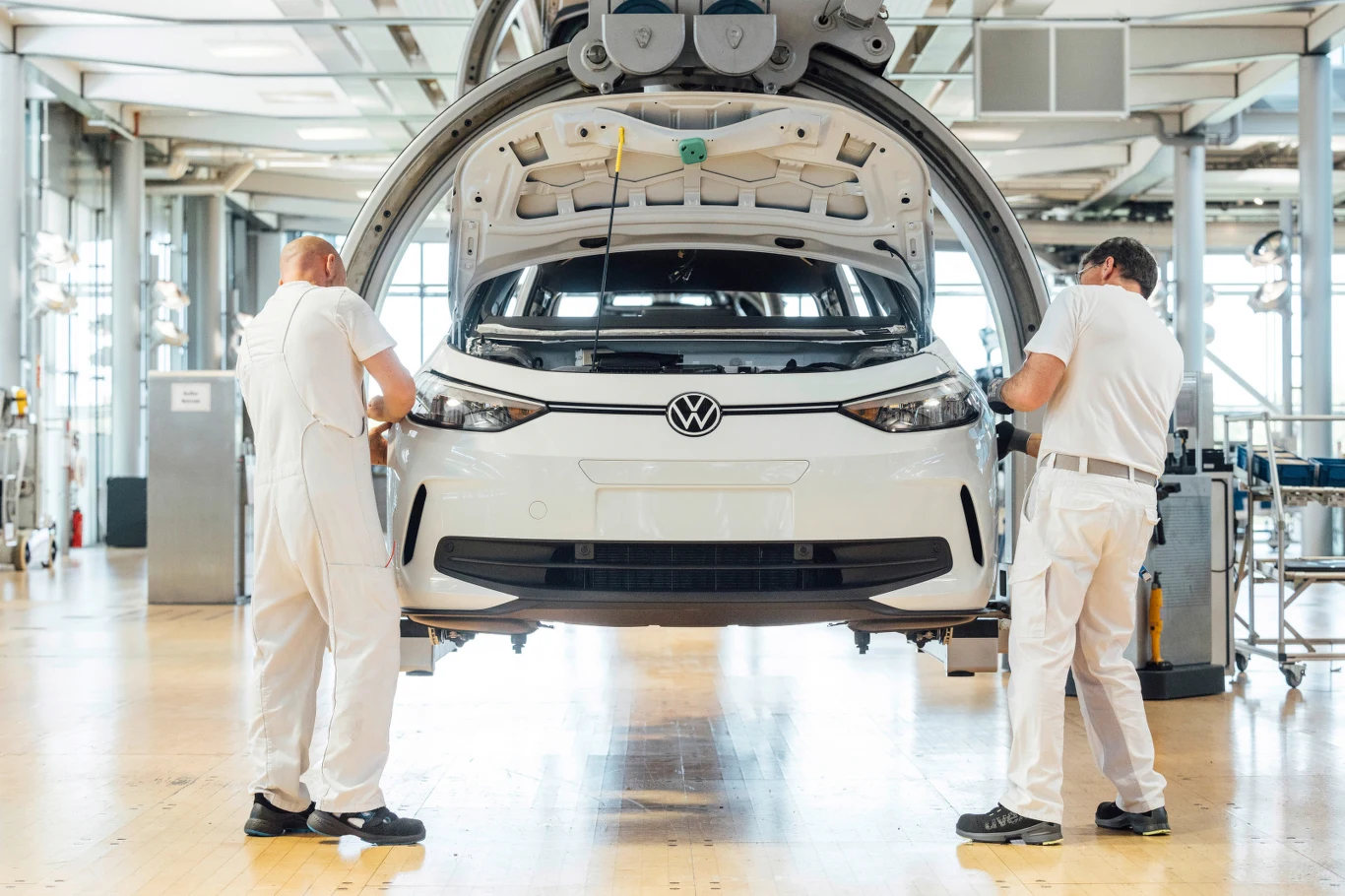 Dwóch pracowników w białych kombinezonach montuje biały samochód marki Volkswagen w nowoczesnej fabryce, jasne wnętrze hali produkcyjnej otacza precyzyjny proces montażu pojazdu.