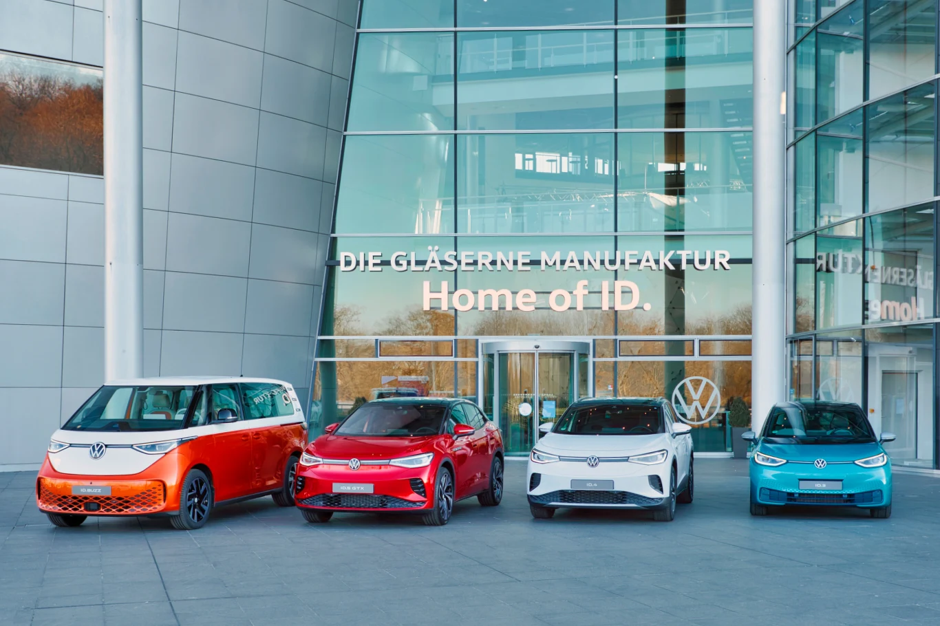 Cztery nowoczesne samochody elektryczne marki Volkswagen w różnych kolorach stoją przed przeszklonym budynkiem z napisem 'Die Gläserne Manufaktur Home of ID.' Budynek oraz logo Volkswagena widoczne w tle.