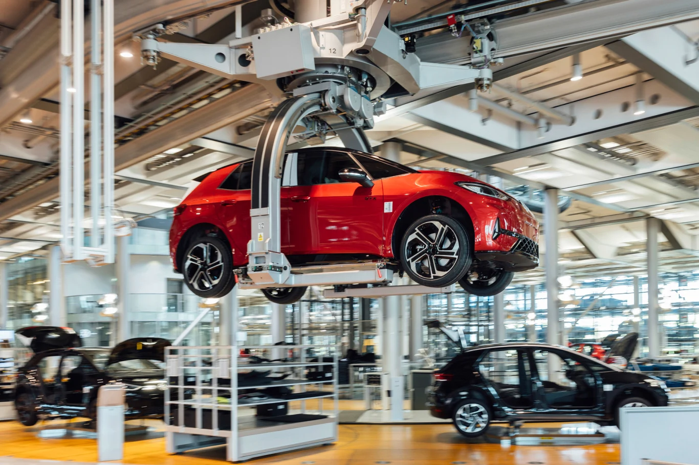 Czerwony samochód elektryczny zawieszony na mechanicznym ramieniu transportującym go przez nowoczesną halę montażową, w tle widoczne inne auta i częściowo zautomatyzowana linia produkcyjna.