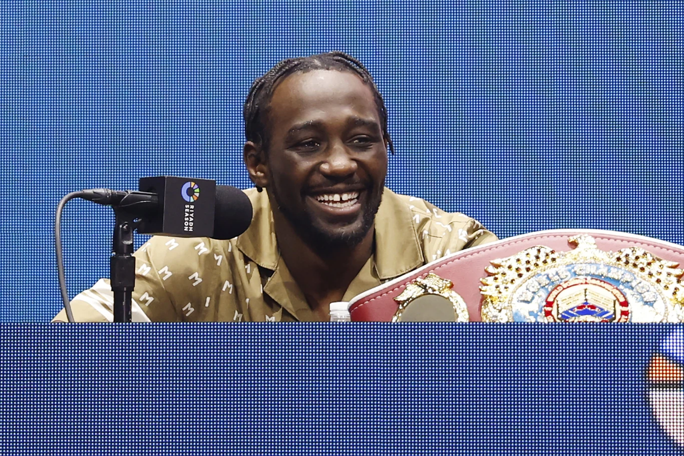 Uśmiechnięty mężczyzna siedzi za stołem konferencyjnym z mikrofonem, a przed nim leży mistrzowski pas bokserski. W tle znajduje się niebieskie, podświetlane tło.