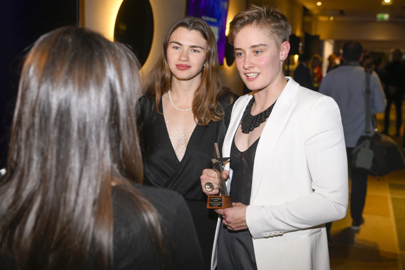 Dwie elegancko ubrane kobiety rozmawiają z trzecią osobą, jedna z nich trzyma w ręku statuetkę, w tle widoczni są inni uczestnicy wydarzenia.
