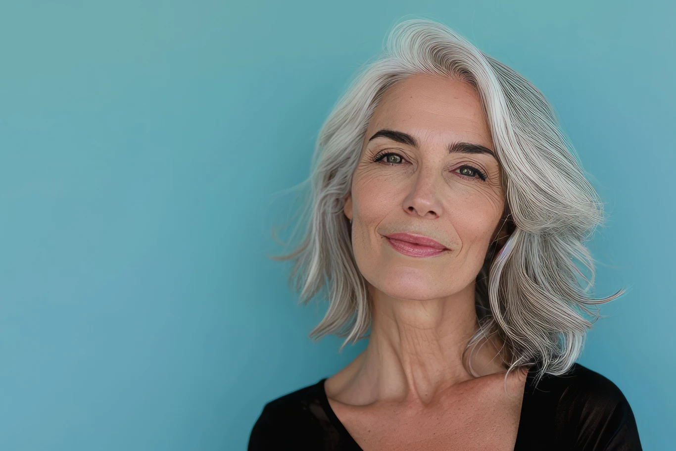 Doświadczona kobieta z siwymi włosami i delikatnym makijażem patrzy spokojnie w stronę aparatu, tło jednolite, turkusowe, ubiór elegancki i klasyczny.