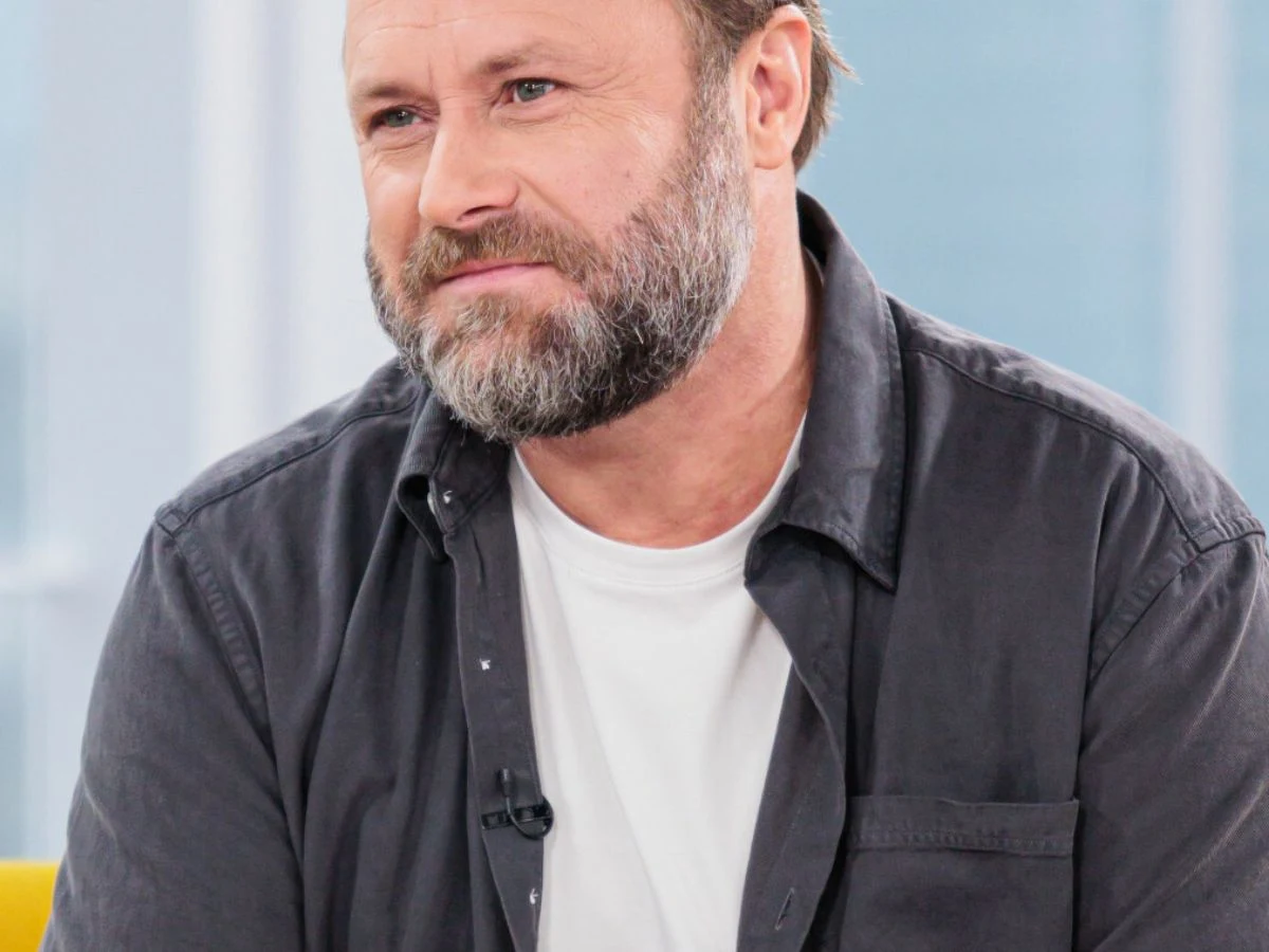 Leszek Lichota Mężczyzna z brodatą twarzą, ubrany w ciemną koszulę i biały t-shirt, siedzi na tle jasnego, rozmytego wnętrza.