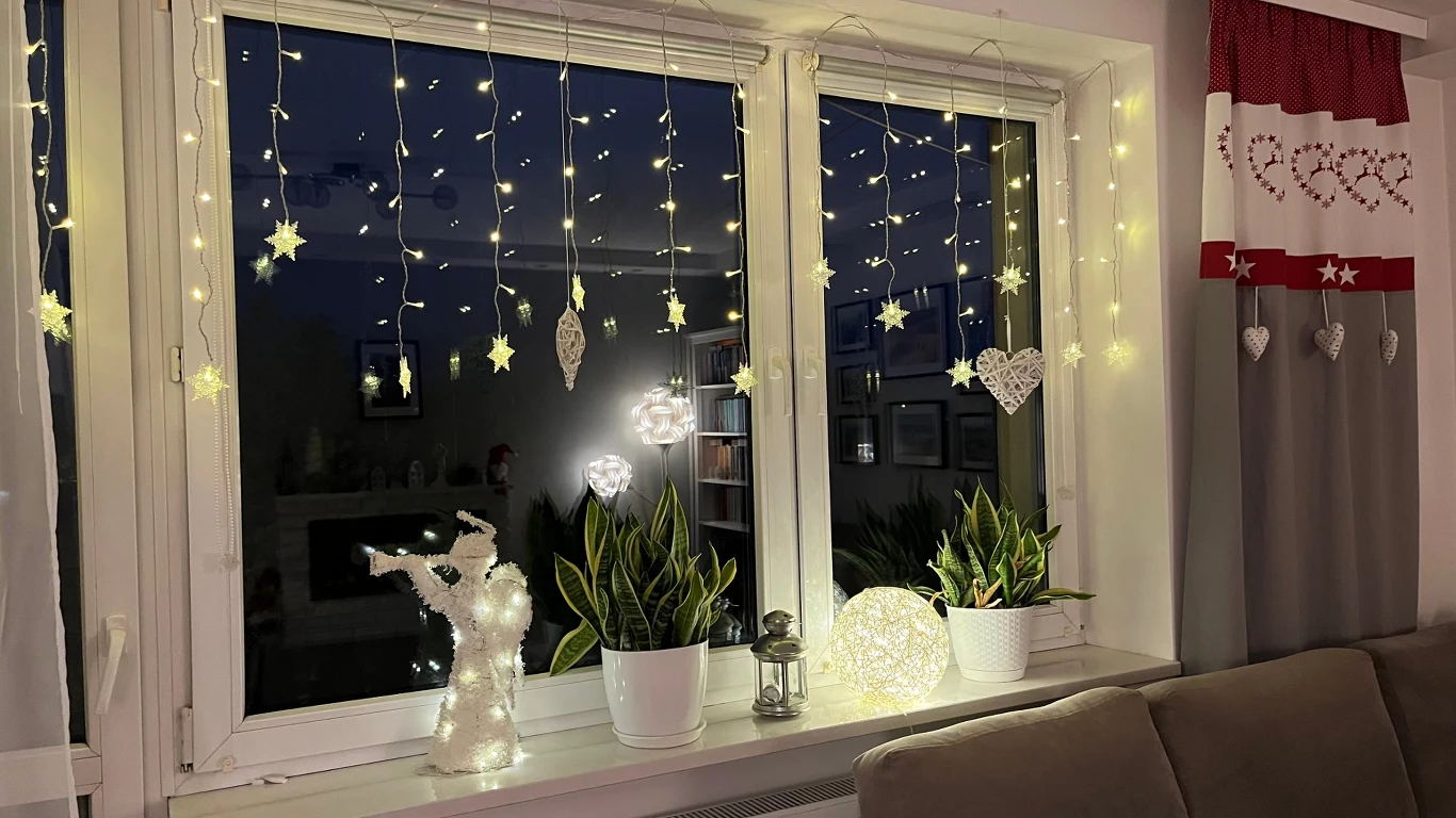 Ozdobione światełkami okno z żywymi roślinami doniczkowymi, świątecznymi zawieszkami w kształcie gwiazdek i serc oraz dekoracyjną figurką anioła i kulą świetlną, tworzące przytulny, świąteczny klimat.