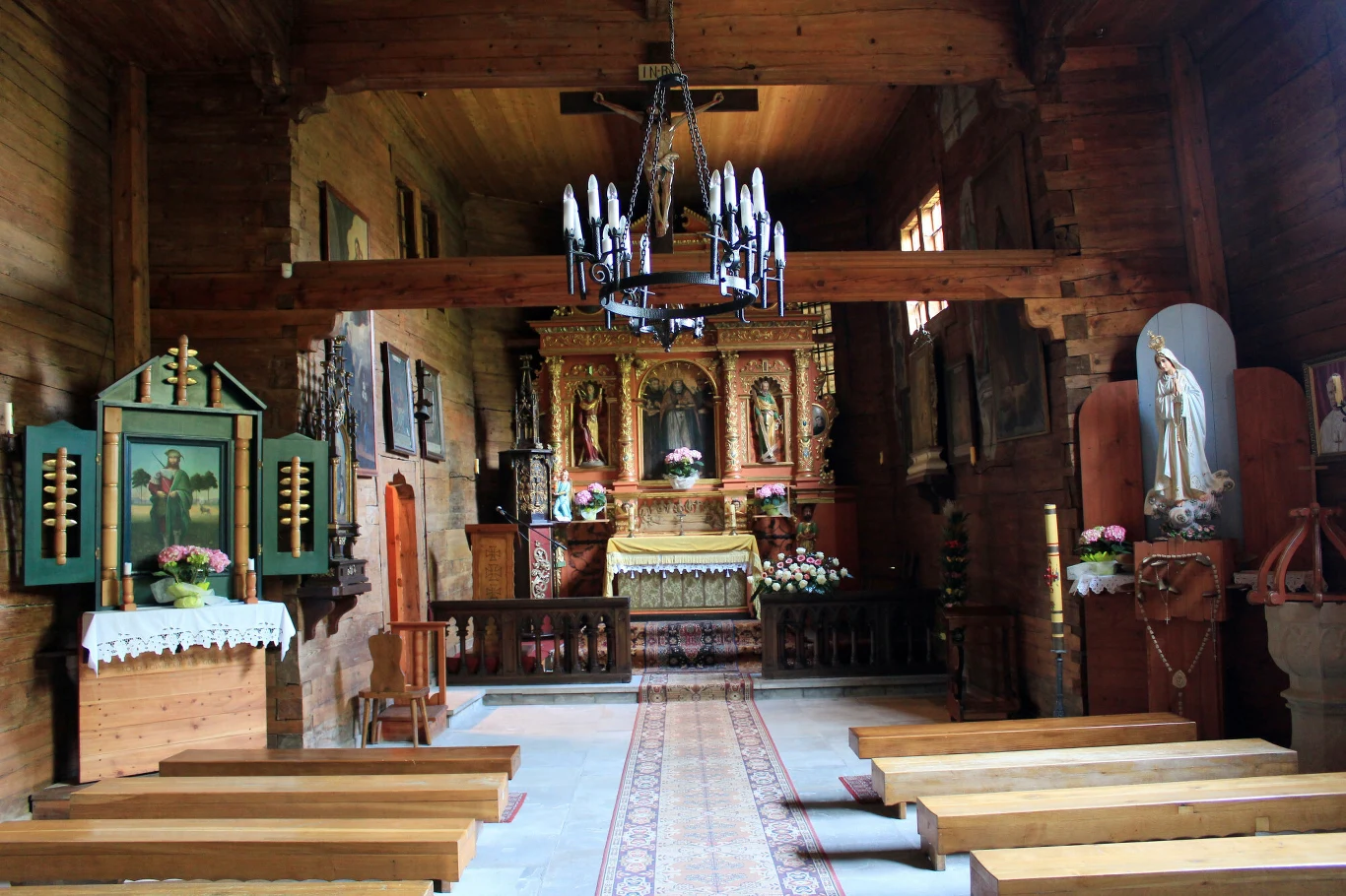 Wnętrze drewnianego kościoła z drewnianymi ławkami, ołtarzem ozdobionym kwiatami oraz figurą Maryi ustawioną po prawej stronie. Na suficie wiszący żyrandol z licznymi świecami, a po bokach zdobione obrazy i tablice.