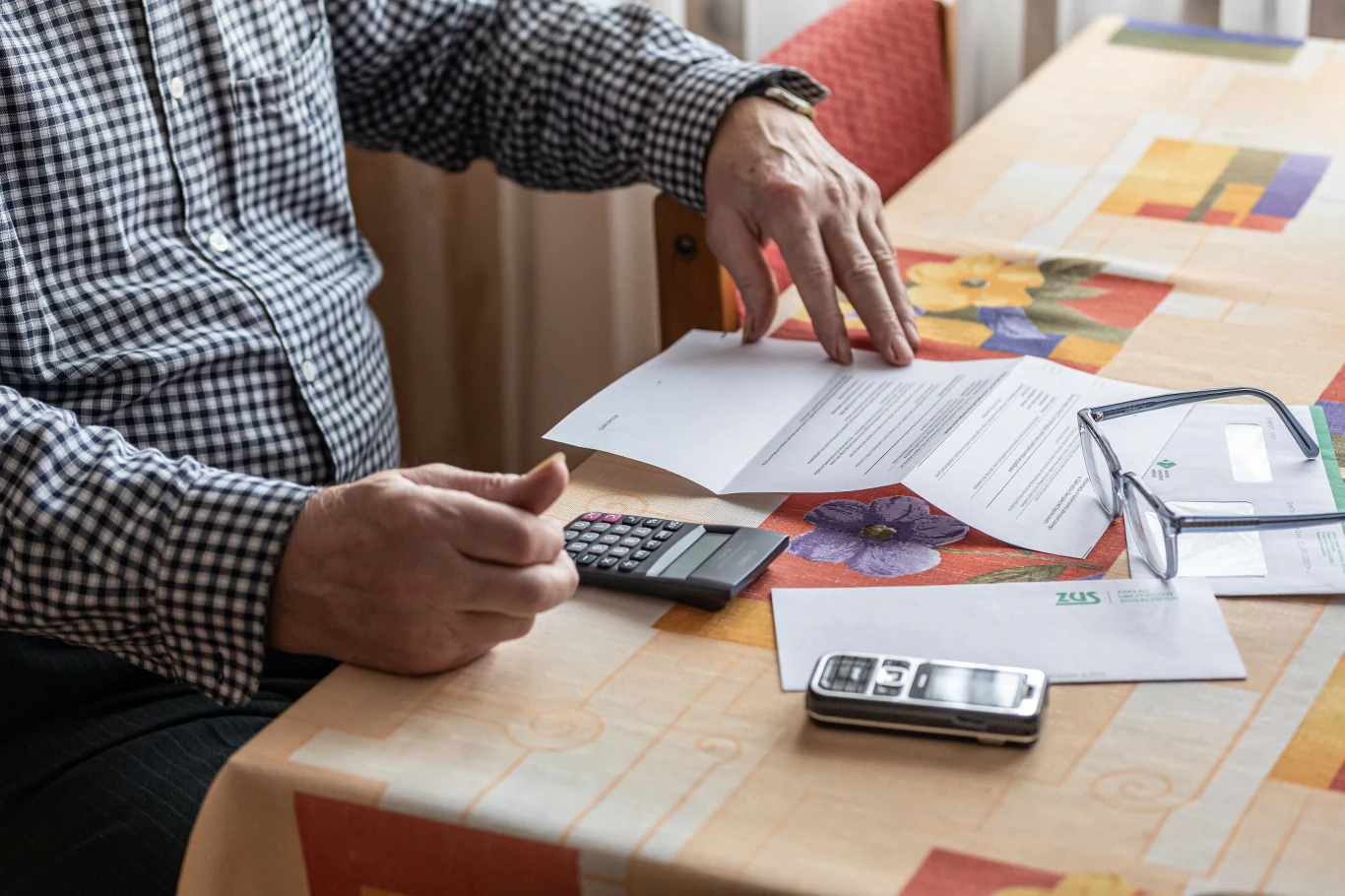 Część emerytów będzie miała wyższe świadczenie. Wystarczy tylko poczekać Dłonie osoby siedzącej przy stole z kolorowym obrusem, korzystającej z kalkulatora i przeglądającej dokumenty, obok leżą okulary i telefon komórkowy.
