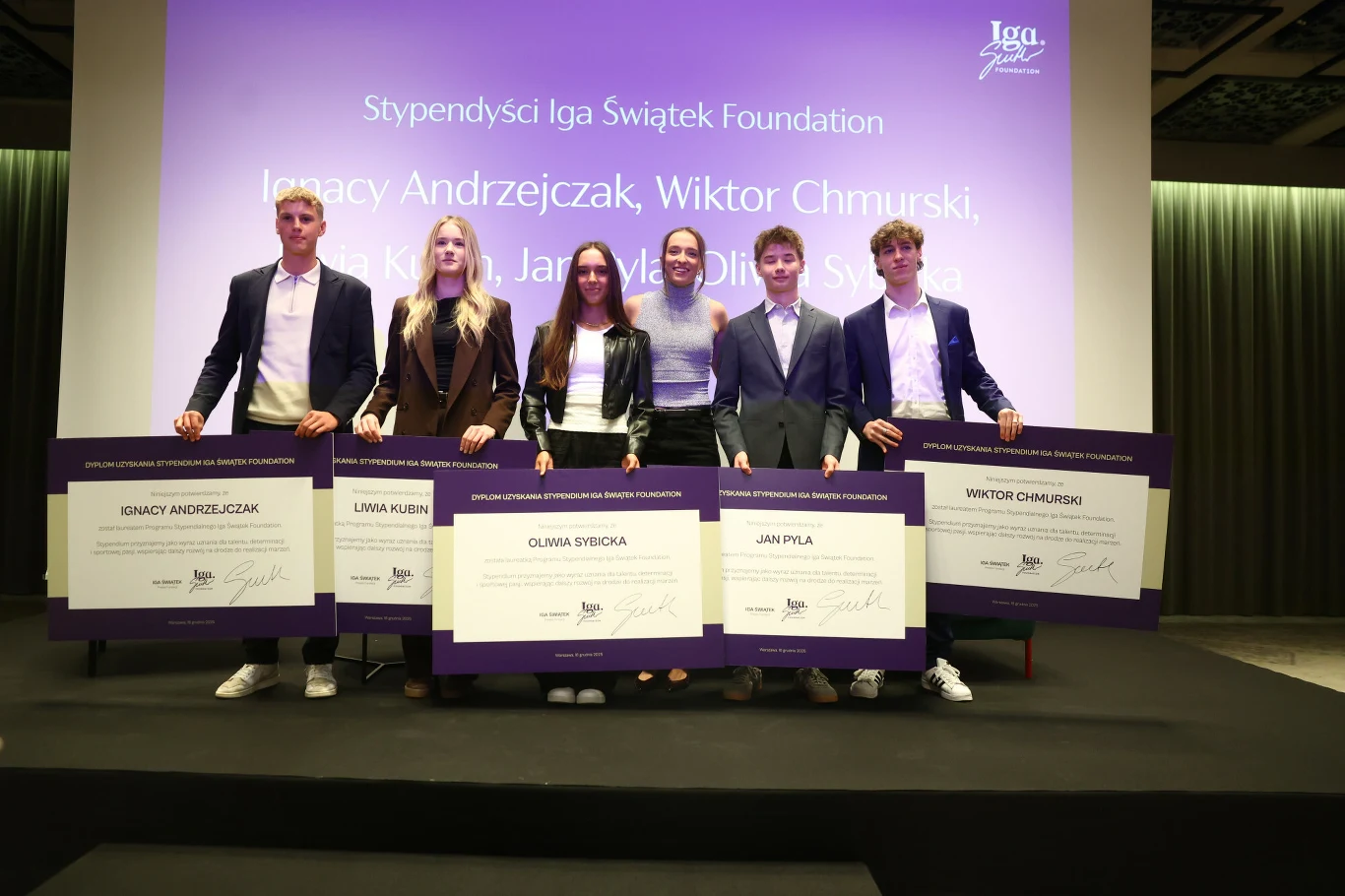 Iga świątek ze stypendystami Grupa młodych ludzi ubrana elegancko trzyma przed sobą duże plansze z informacjami o stypendiach. W tle wyświetlona jest nazwa fundacji oraz imiona i nazwiska stypendystów.