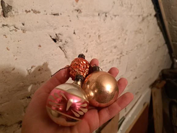 Trzy bombki choinkowe w różnych kolorach i wzorach trzymane w dłoni na tle białej, nierównej ściany.