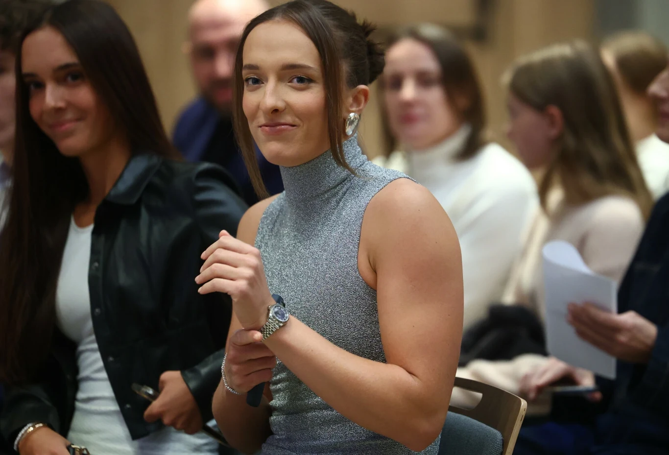 Iga Świątek ogłosiła ważne nowiny Młoda kobieta siedząca na krześle, ubrana w srebrny bezrękawnik, uśmiecha się subtelnie patrząc w stronę obiektywu. W tle kilka innych osób, które rozmawiają lub trzymają kartki papieru, atmosfera przypomina wydarzenie oficjalne lub spotkanie.