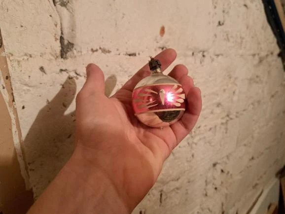 Dłoń trzyma kolorową, błyszczącą bombkę choinkową z wzorem w odcieniach różu i srebra na tle białej, ceglanej ściany.