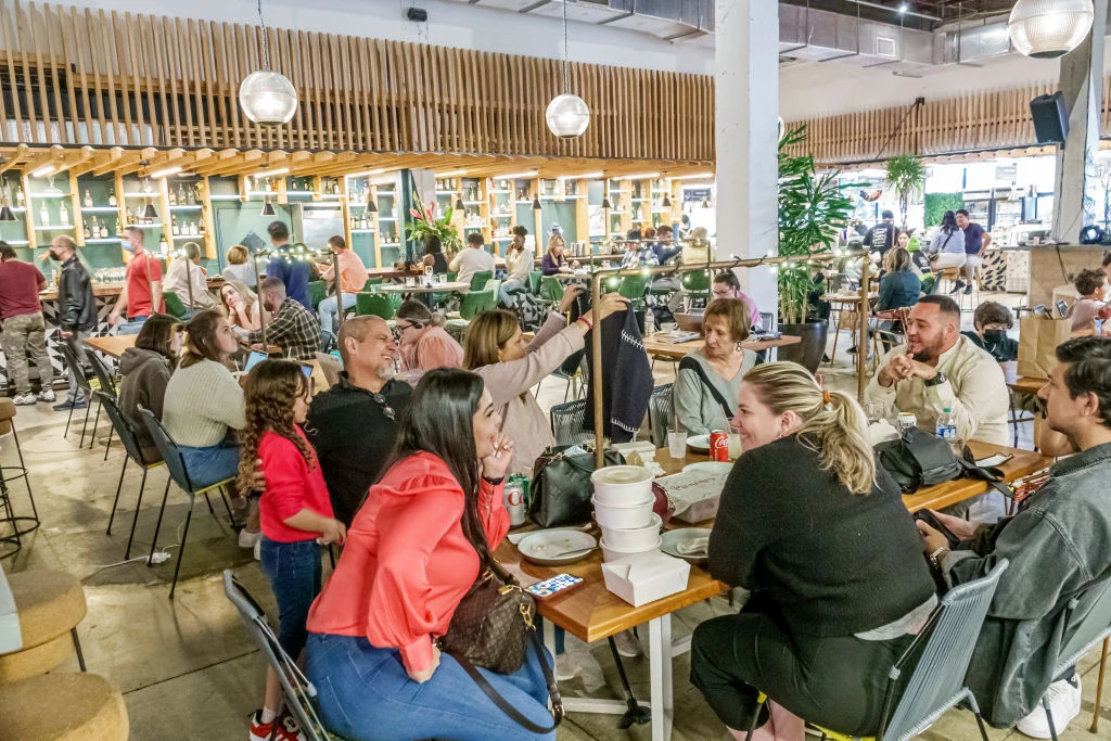 Ludzie siedzący przy stołach w nowoczesnej, tętniącej życiem restauracji, rozmawiają i jedzą, wokół widać jasne oświetlenie oraz drewniane i zielone akcenty wystroju wnętrza.