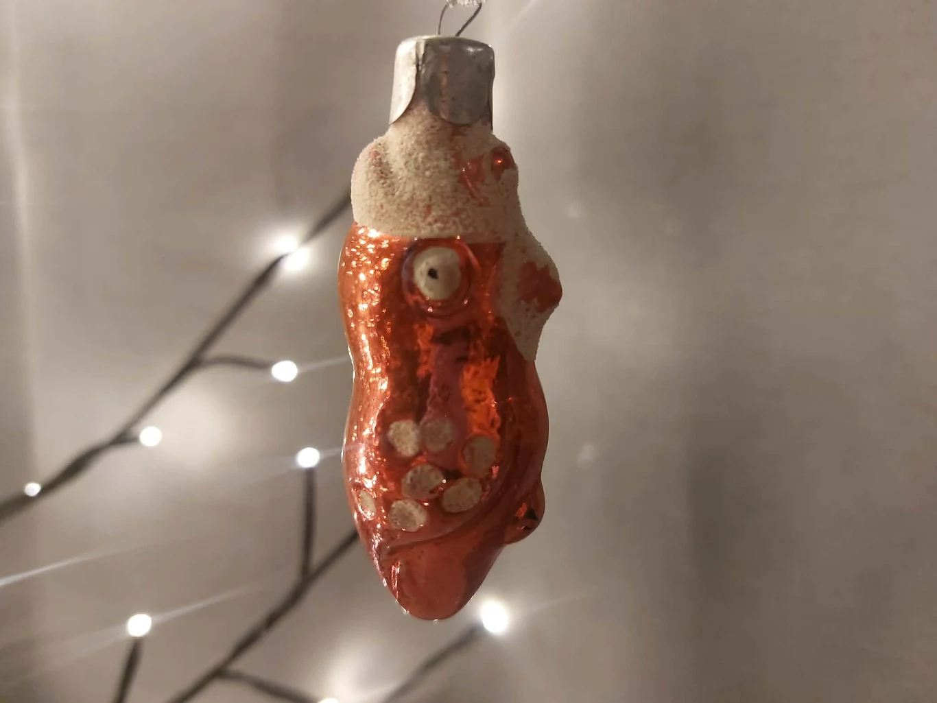 Ozdoba choinkowa o błyszczącej, czerwonej powierzchni, pokryta dekoracyjnymi kropkami i niewyraźnym wzorem, zawieszona na tle rozświetlonych lampek choinkowych.