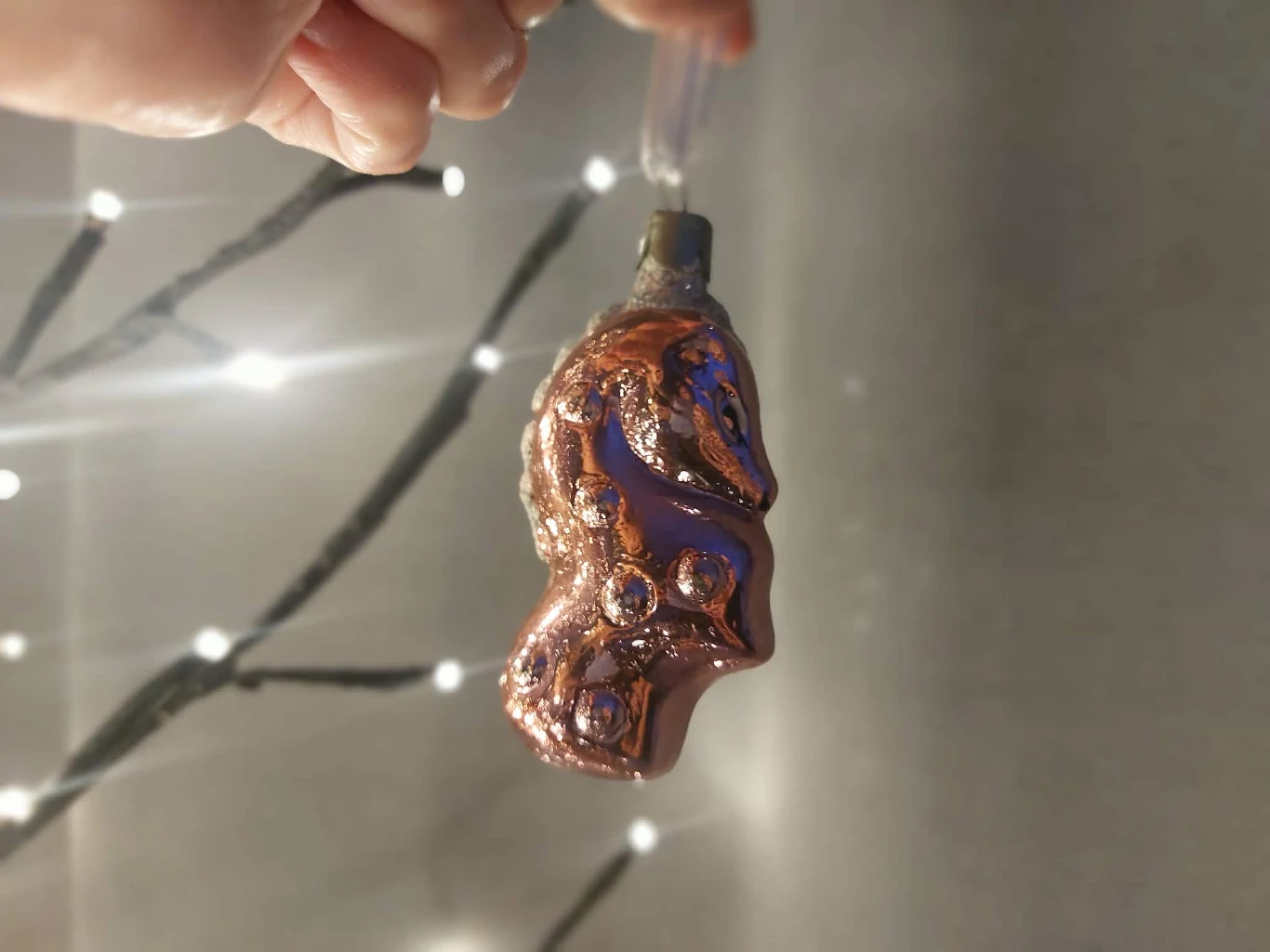 Szklana bombka choinkowa w kształcie ozdobnej figurki trzymana za zawieszkę, na tle rozświetlonych światełek dekoracyjnych.