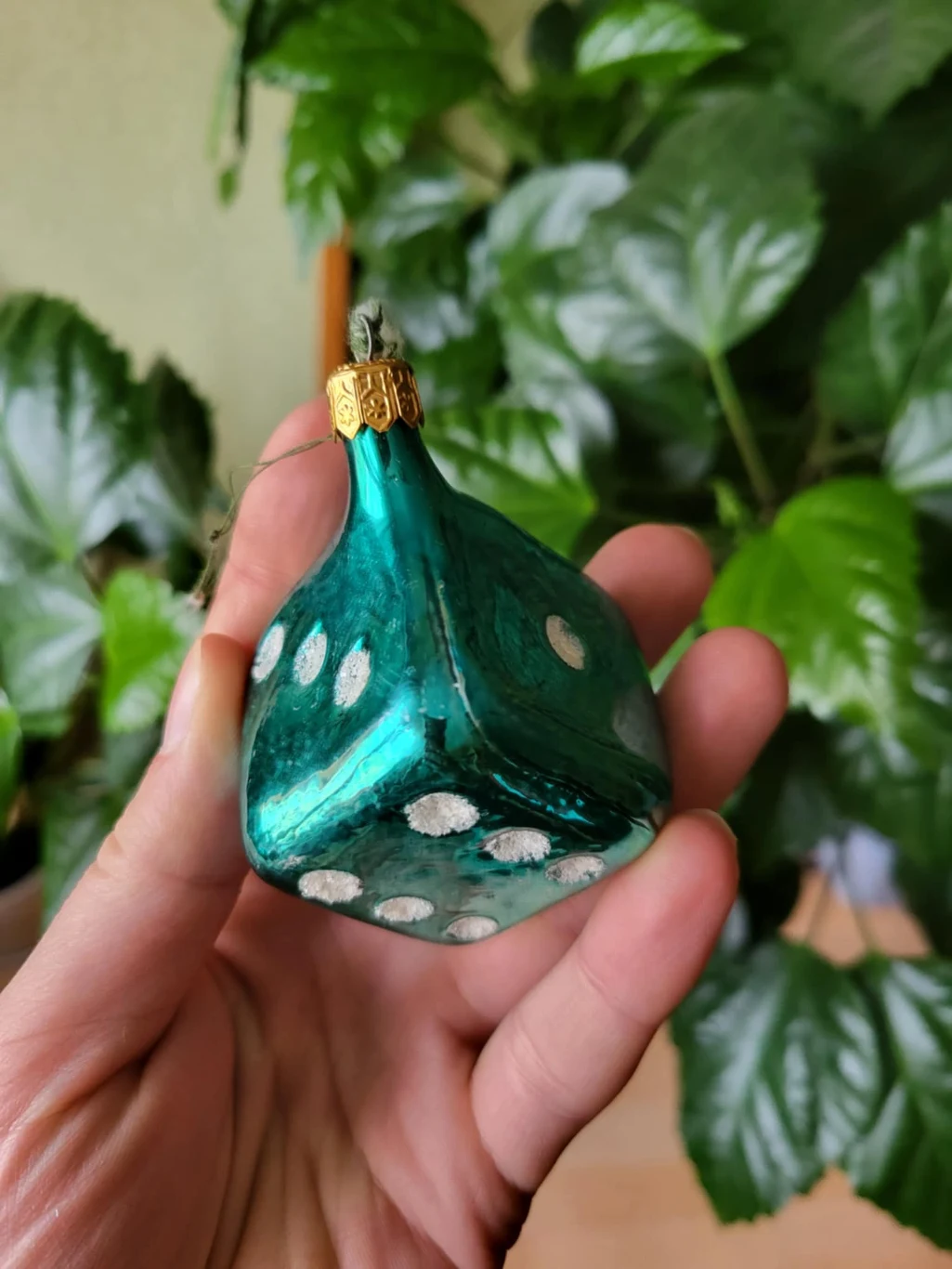 Szklana bombka w kształcie kostki do gry w zielonym kolorze z białymi kropkami, trzymana w dłoni na tle zielonych liści rośliny doniczkowej.