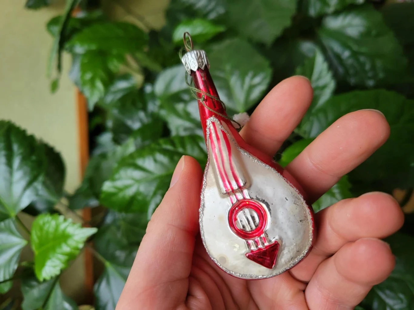 Szklana ozdoba choinkowa w kształcie mandoliny trzymana w dłoni, w tle zielone liście rośliny doniczkowej.