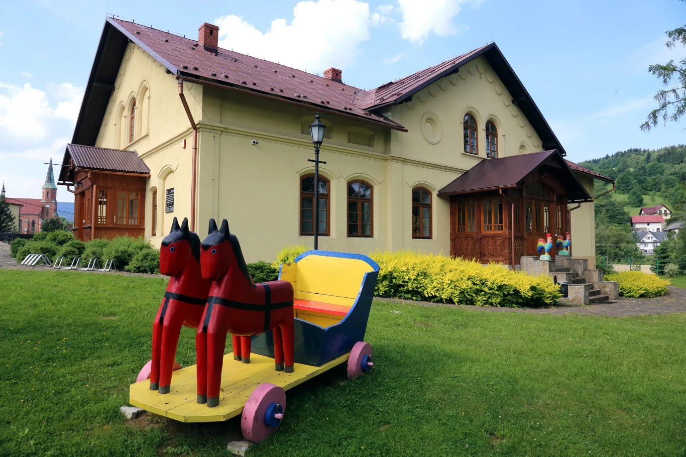 Duży żółty budynek z czerwonym dachem otoczony zielonym trawnikiem i krzewami, na pierwszym planie kolorowa, drewniana rzeźba przedstawiająca dwie czerwone konie ciągnące wózek. W tle widać fragmenty innych domów i niewielkie wzgórza.