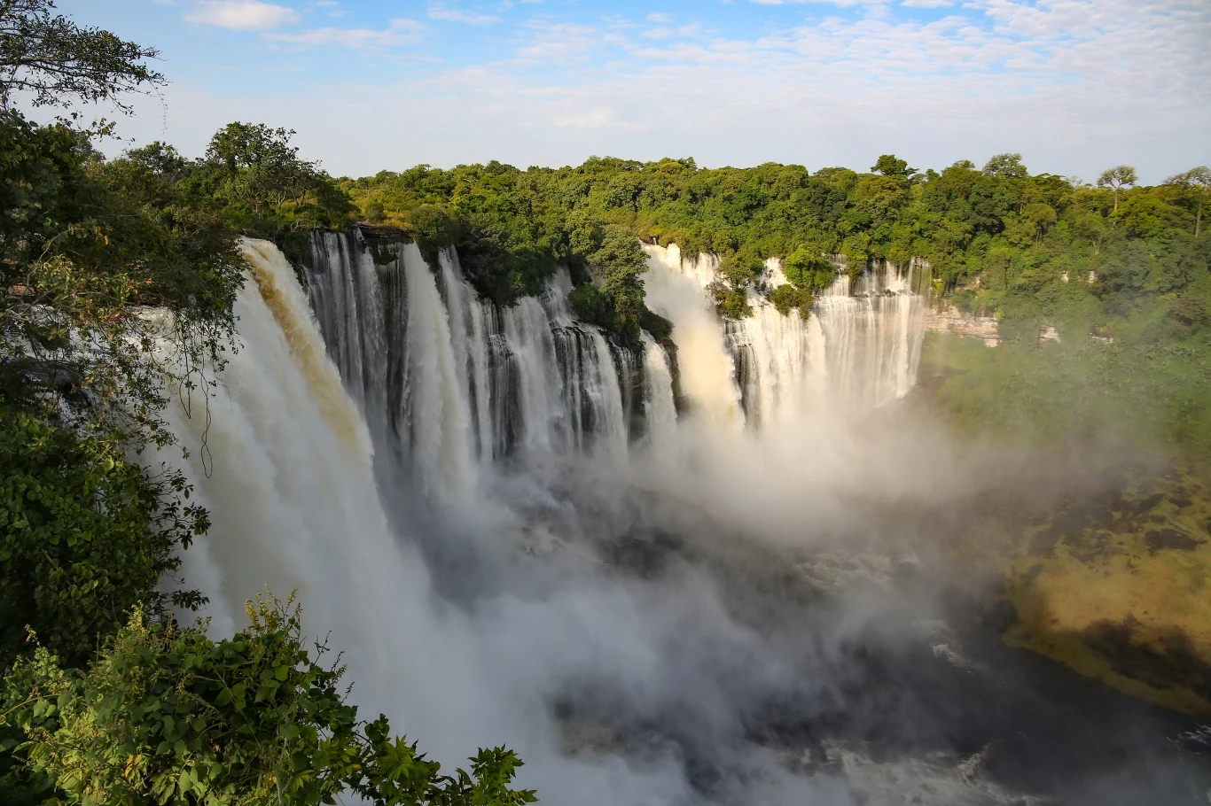Potężny wodospad opadający z wysokiego klifu otoczony gęstą zielenią drzew, z rozpryskującą się wodą i delikatną mgłą tworzącą się przy podnóżu wodospadu.