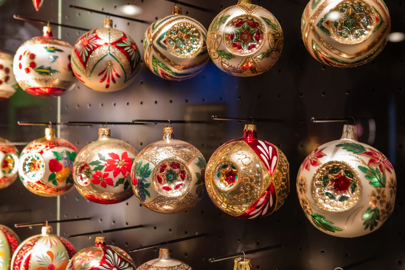 Kolorowe, ozdobne bombki choinkowe wiszące na ekspozycji, zdobione brokatem, motywami świątecznymi i kwiatowymi. W tle połyskujące światła wzmacniają świąteczny klimat.
