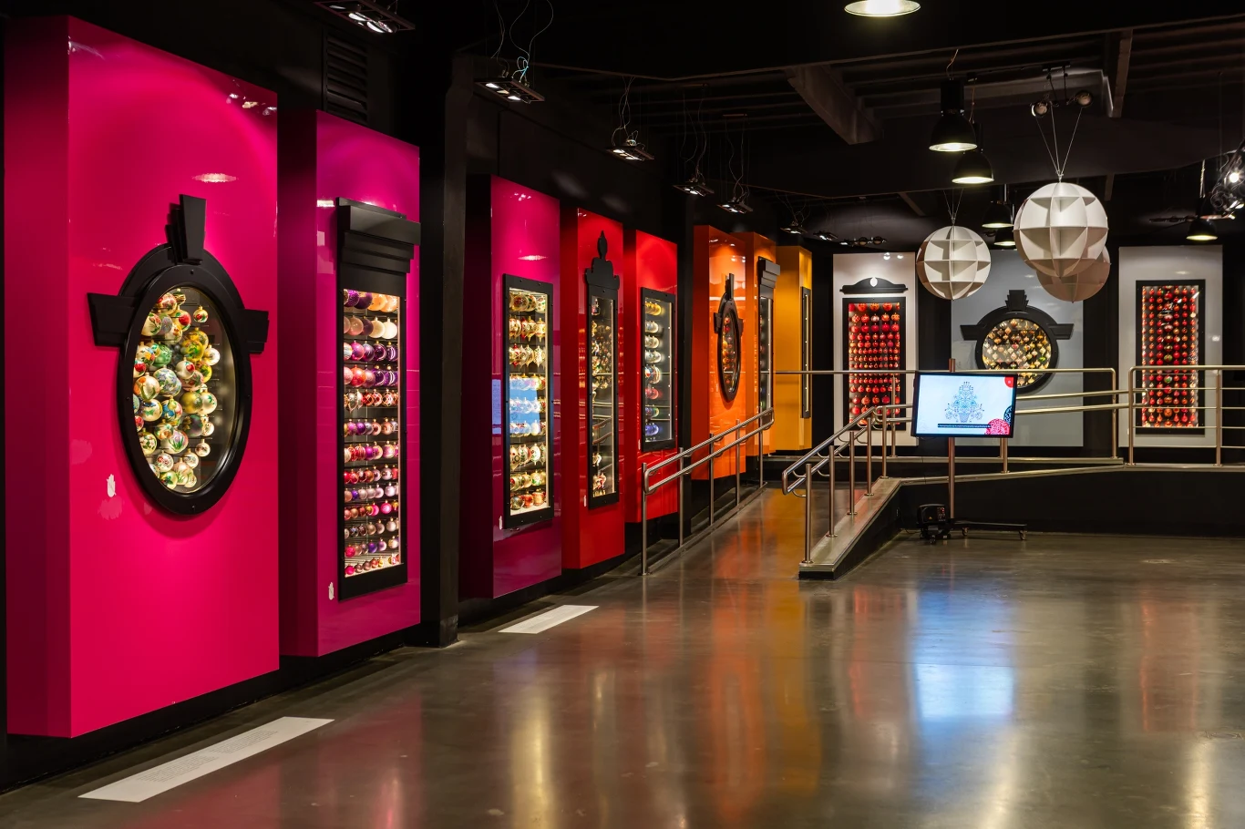 Kolorowe ściany ekspozycyjne z licznymi, podświetlonymi gablotami prezentującymi kolekcje różnorodnych, ozdobnych przedmiotów, rampa prowadząca przez środek pomieszczenia, lśniąca podłoga, oraz nowoczesne dekoracje sufitowe.