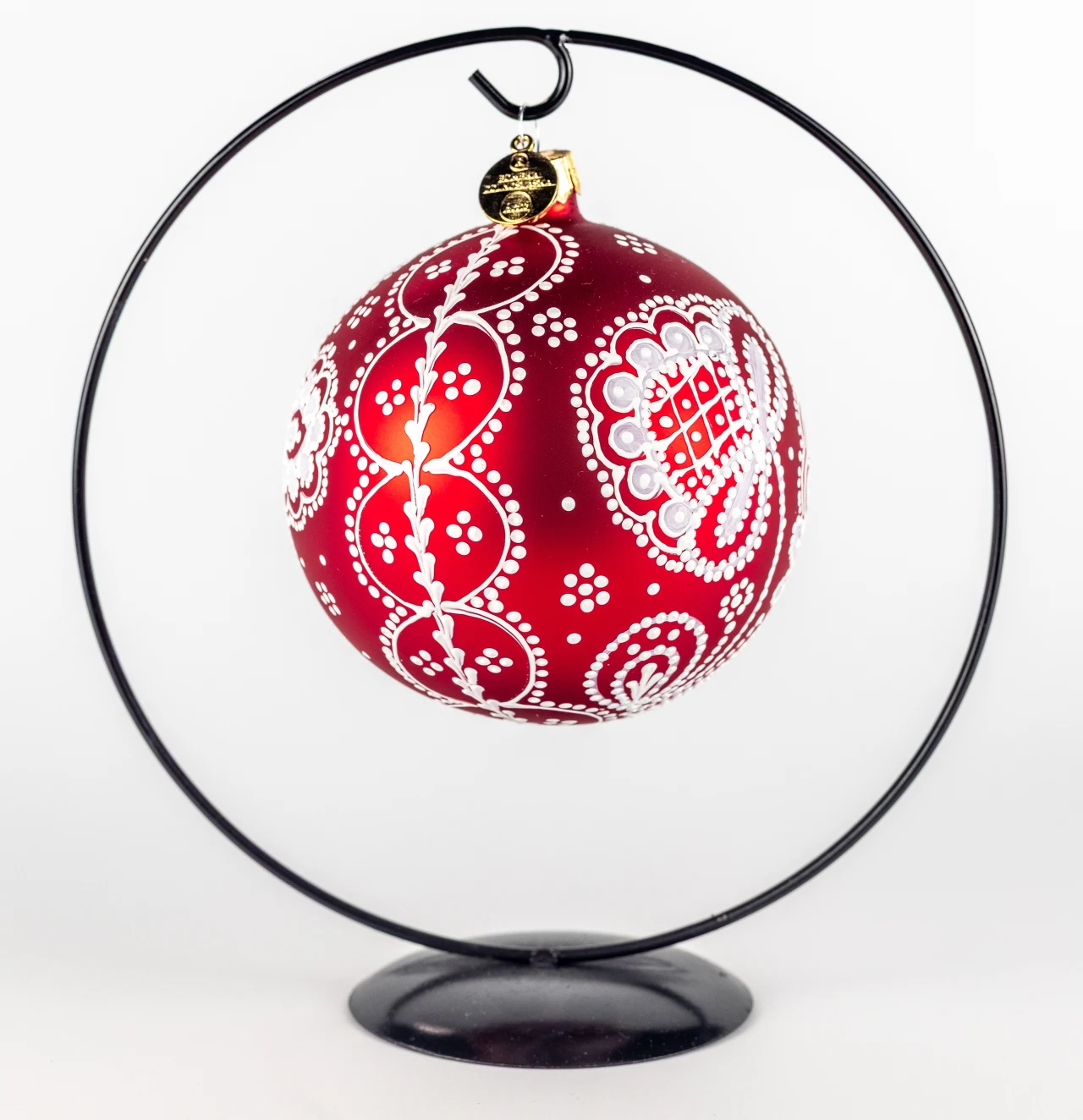 Czerwona bombka choinkowa ozdobiona białym, koronkowym wzorem, zawieszona na metalowym stojaku o okrągłym kształcie, całość na jasnym tle