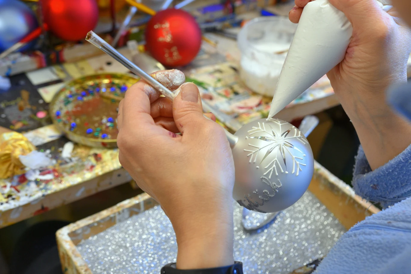 Ręce osoby ozdabiające srebrną bombkę choinkową białą farbą w kształcie śnieżynki, w tle widoczne kolorowe bombki oraz przybory do dekoracji.