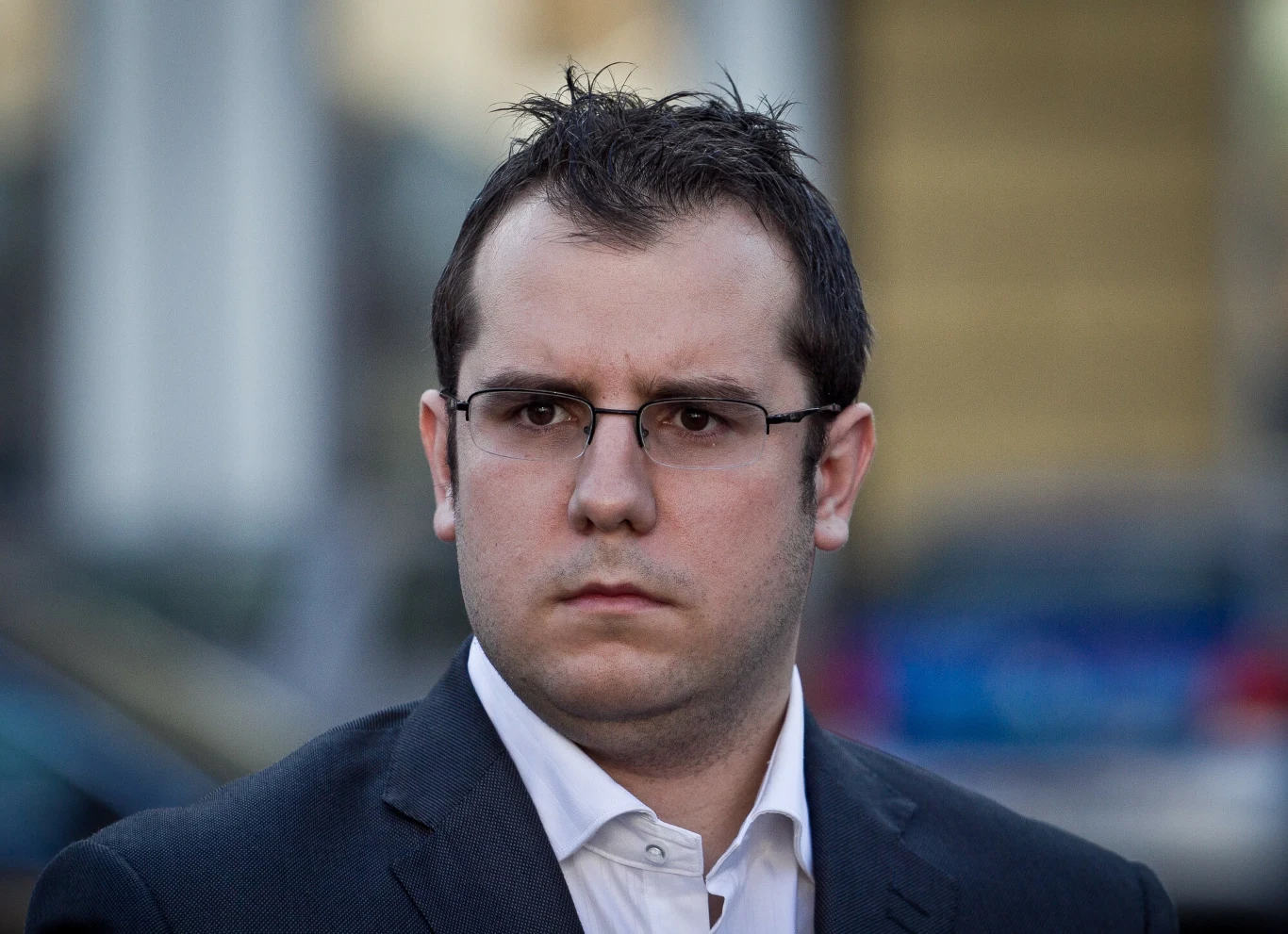 Przemysław Czarnecki Młody mężczyzna w garniturze i okularach patrzy poważnie przed siebie, tło rozmyte z widocznymi budynkami i samochodami.