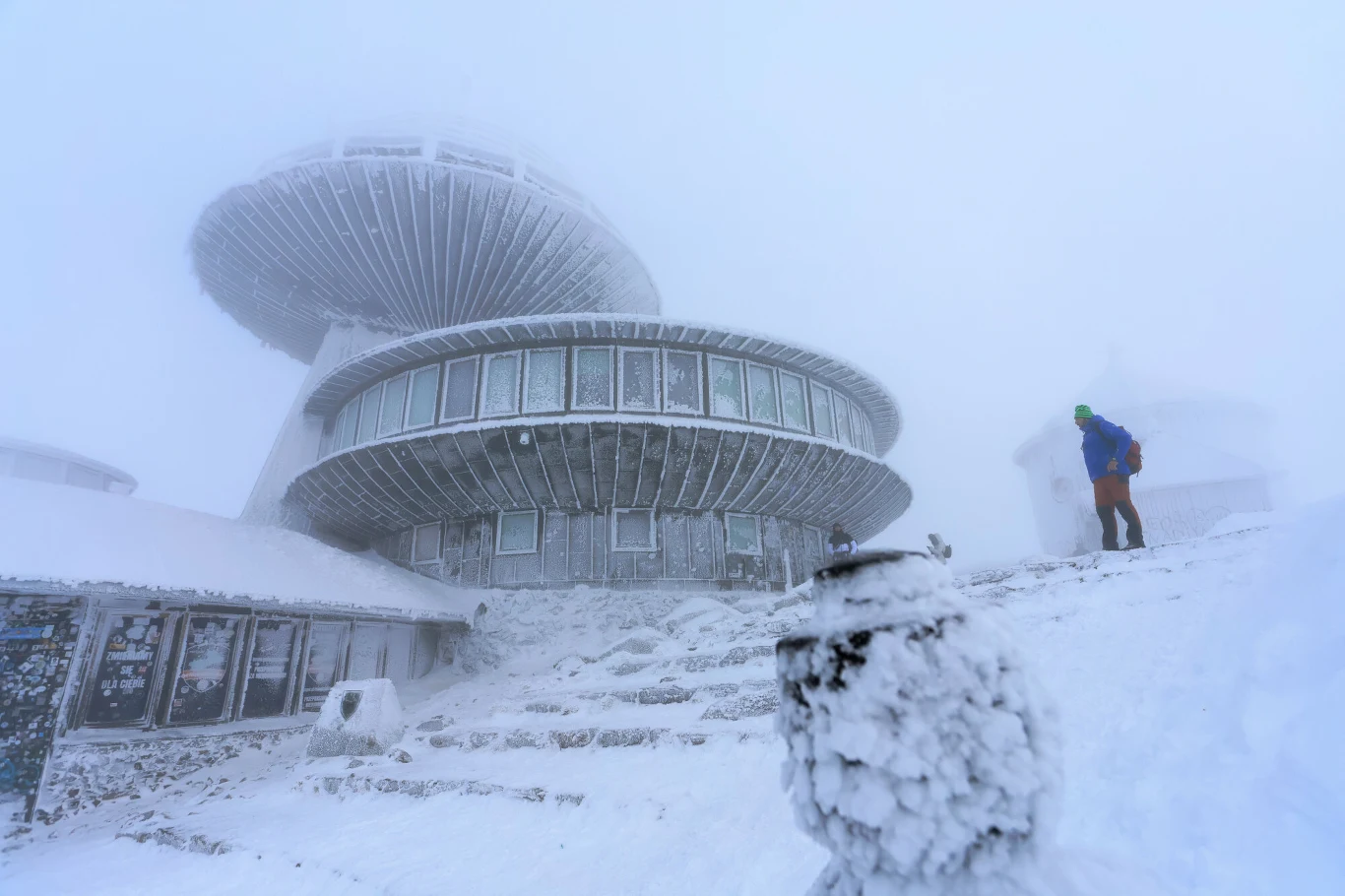 Modernistyczny budynek naśnieżony i oszroniony, przypominający wyglądem spodki UFO, na tle intensywnej zimowej mgły. Na schodach pokrytych śniegiem stoi ubrana w niebieską kurtkę i zieloną czapkę osoba, w oddali niewyraźnie widoczna sylwetka innej kons...
