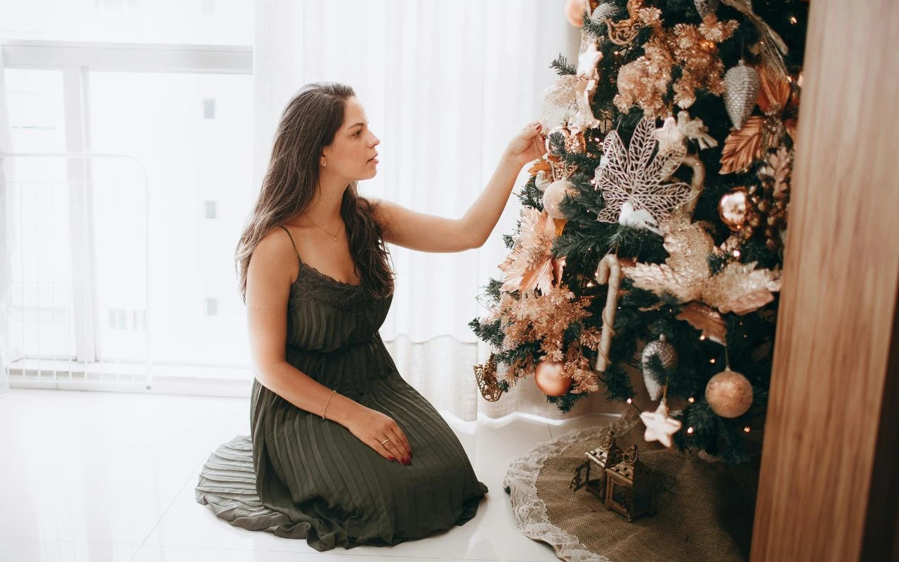 Kobieta w ciemnozielonej sukience klęczy obok bogato udekorowanej choinki, skupiając się na zawieszaniu ozdoby. W tle jasne zasłony i wnętrze mieszkania, wokół drzewka widoczne prezenty i świąteczne dekoracje.