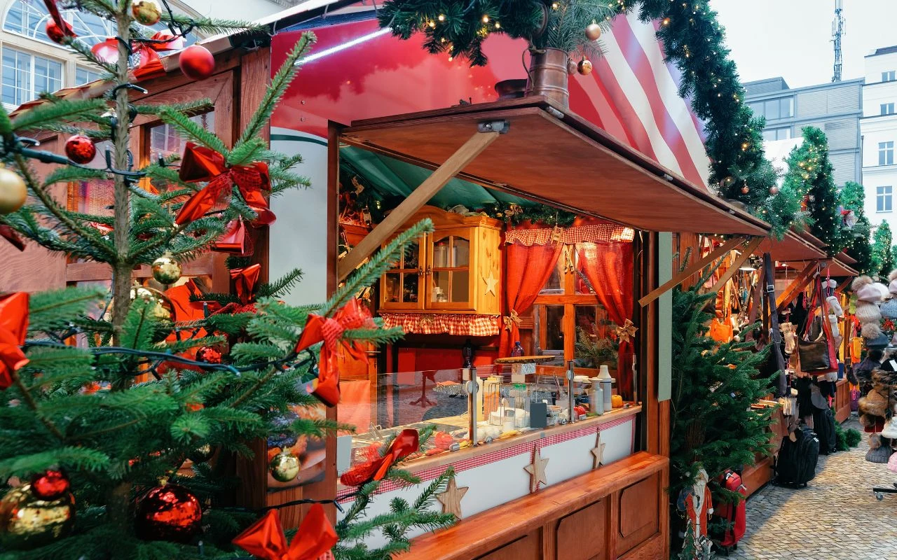 Stoisko na jarmarku bożonarodzeniowym ozdobione zielonymi gałązkami i czerwonymi kokardami, pełne świątecznych produktów i upominków, w tle choinki oraz fragmenty ludzi odwiedzających targ.