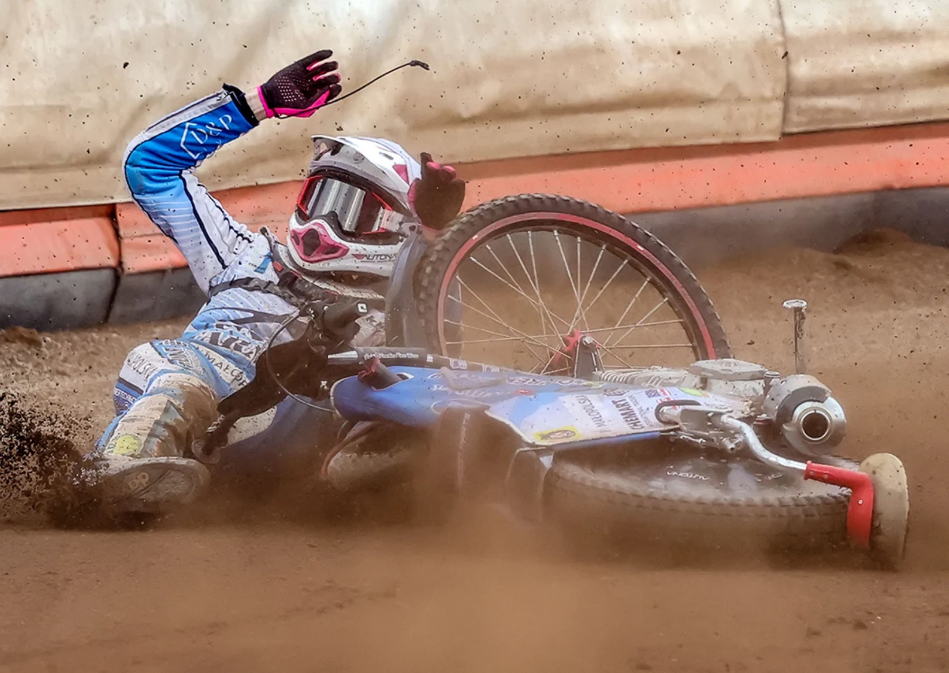 Zawodnik sportów motorowych leży na torze obok przewróconego motocykla, unosząca się wokół kurz podkreśla dynamiczność i gwałtowność upadku, na zawodniku pełny strój ochronny oraz kask.