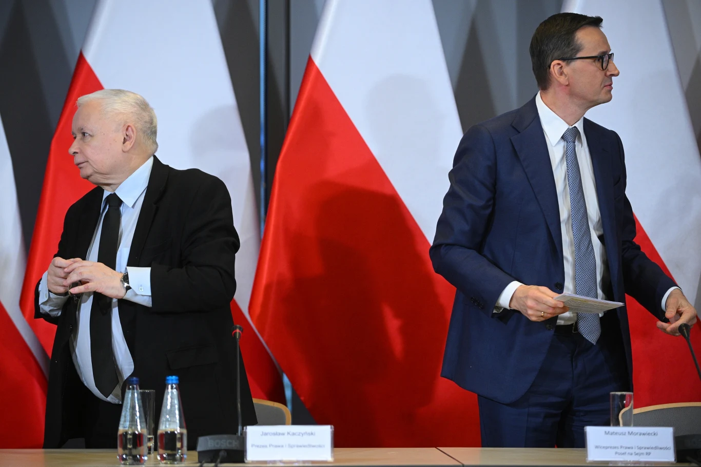 Jarosław Kaczyński i Mateusz Morawiecki Dwóch mężczyzn w garniturach stoi przy stole konferencyjnym, za nimi biało-czerwone flagi Polski, na stole widoczne są mikrofony, butelki wody oraz tabliczki z nazwiskami.