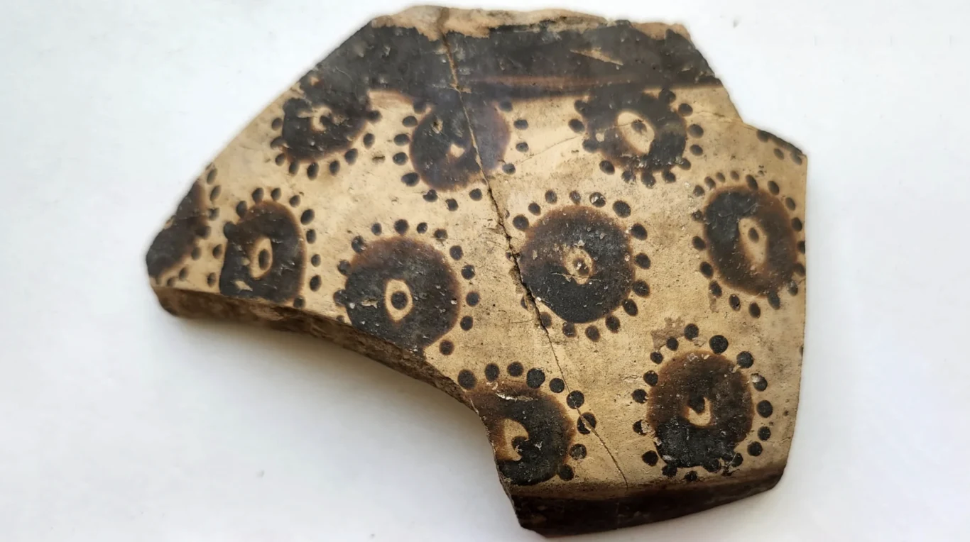 Nax Mallowan excavation at Arpachiyah, Iraq. From the collections of the British Museum and UCL. Credit: Yosef Garfinkel Fragment ceramiki o jasnym tle, ozdobiony ciemnymi okręgami z kropkami ułożonymi wokół każdego z nich, świadczący o dawnej technice zdobienia naczyń.