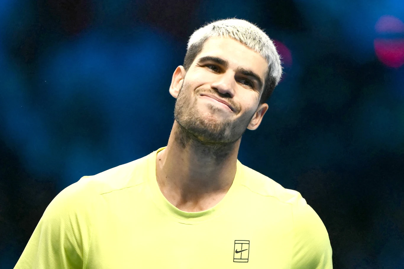 Młody mężczyzna o jasnych, krótkich włosach ubrany w żółtą koszulkę sportową stoi na tle rozmytego, niebieskiego tła, z lekkim wyrazem niezadowolenia na twarzy.