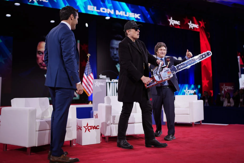 Trzech mężczyzn stoi na scenie podczas konferencji, środkowa postać trzyma dużą piłę łańcuchową, w tle znajdują się ekrany z napisem Elon Musk i logotypem CPAC, na scenie ustawione są białe fotele oraz amerykańska flaga.